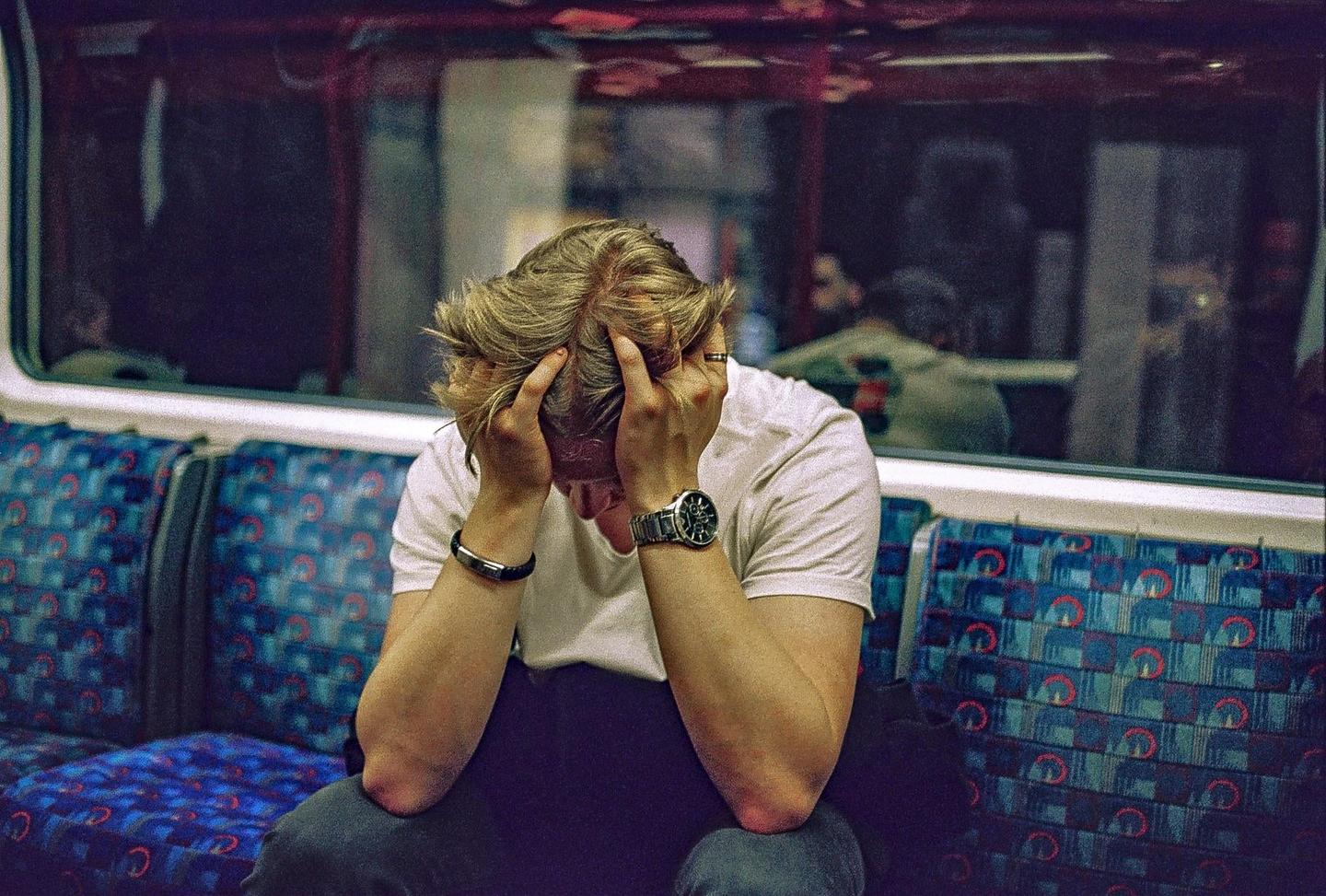 A man sits on a train with his head in his hands, looking distressed.