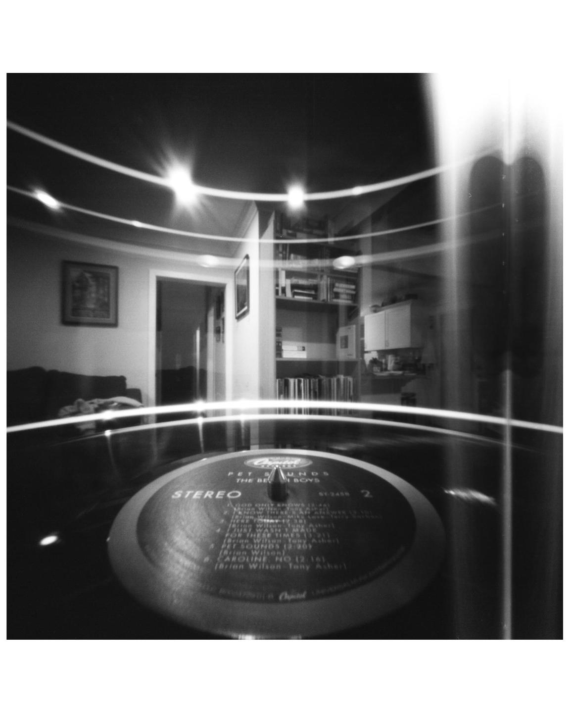 A record player with a vinyl record is in the foreground, with a room and bookshelves visible in the background.