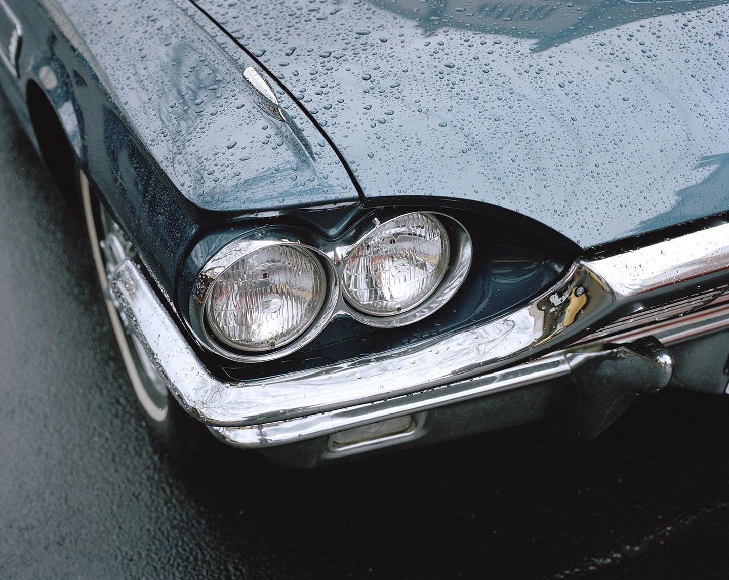 The front of a teal vintage car is covered in raindrops.