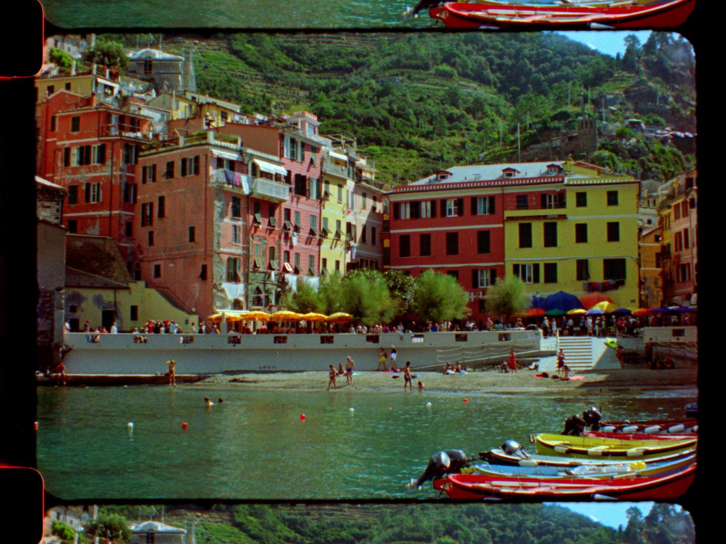 A colorful village with boats in the water and people on the beach is nestled against a green hillside.