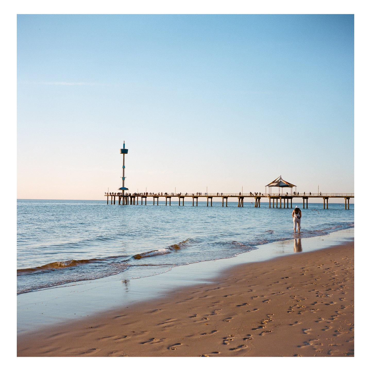 A person stands in the water on a sandy beach with a long pier extending into the ocean under a clear sky.