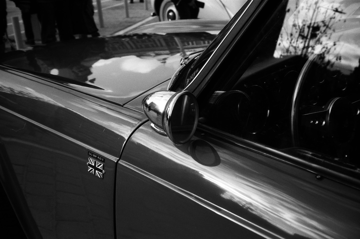 A black and white close-up shows the side of a vintage car with a Union Jack emblem and a chrome side mirror.