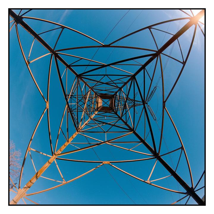 A low-angle view shows the geometric structure of a power line tower against a clear blue sky.