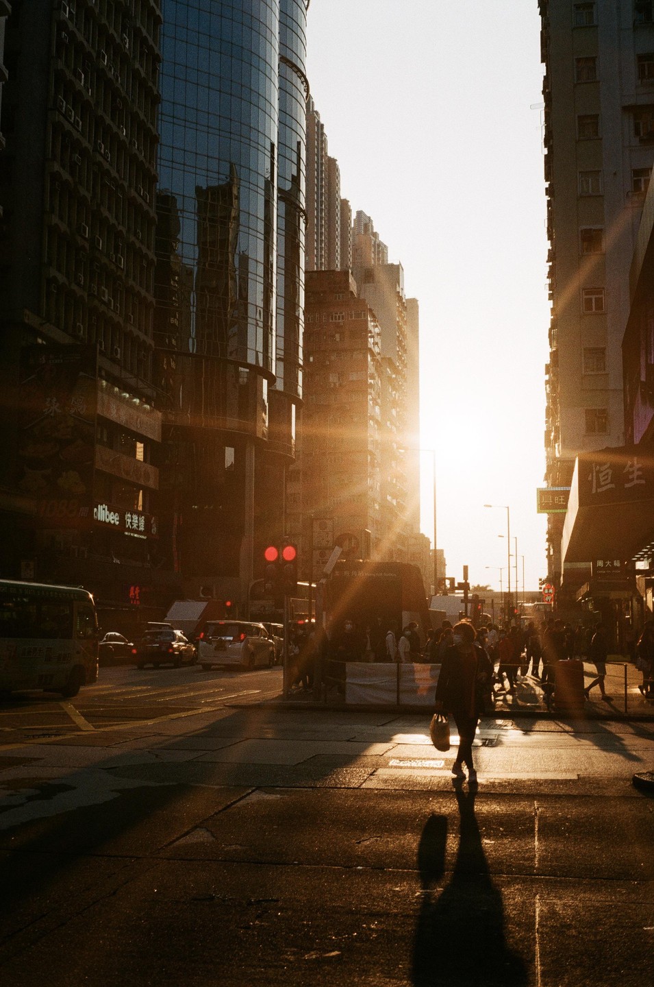The sun sets between tall buildings, casting long shadows on a busy city street with pedestrians and vehicles.