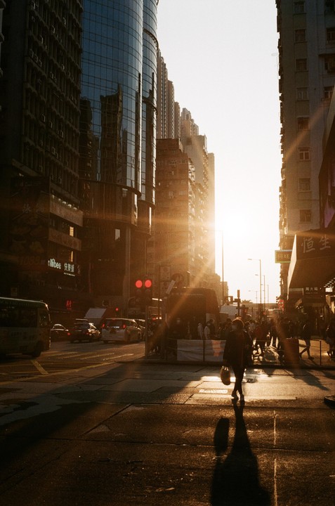 The sun sets between tall buildings, casting long shadows on a busy city street with pedestrians and vehicles.