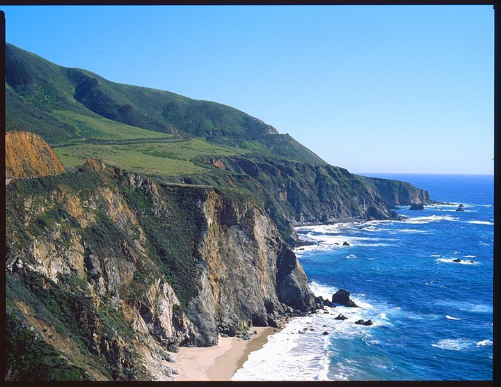 The Pacific Ocean crashes against the rocky Big Sur coastline, with a sandy beach visible at the base of the cliffs.