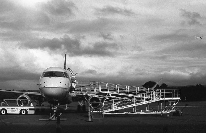 A large airplane is parked on an airfield with a boarding ramp extended to its door, while a smaller plane flies in the distance under a cloudy sky.
