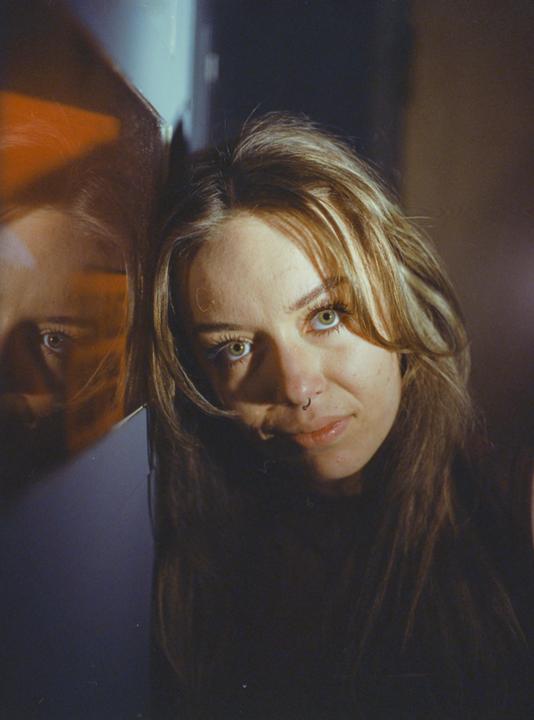 A woman with long hair and a nose ring leans against a reflective surface, with her reflection visible beside her.