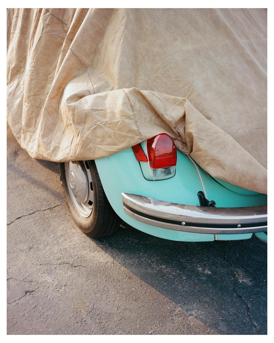 A teal car is partially covered by a tan car cover, revealing a red taillight and chrome bumper.