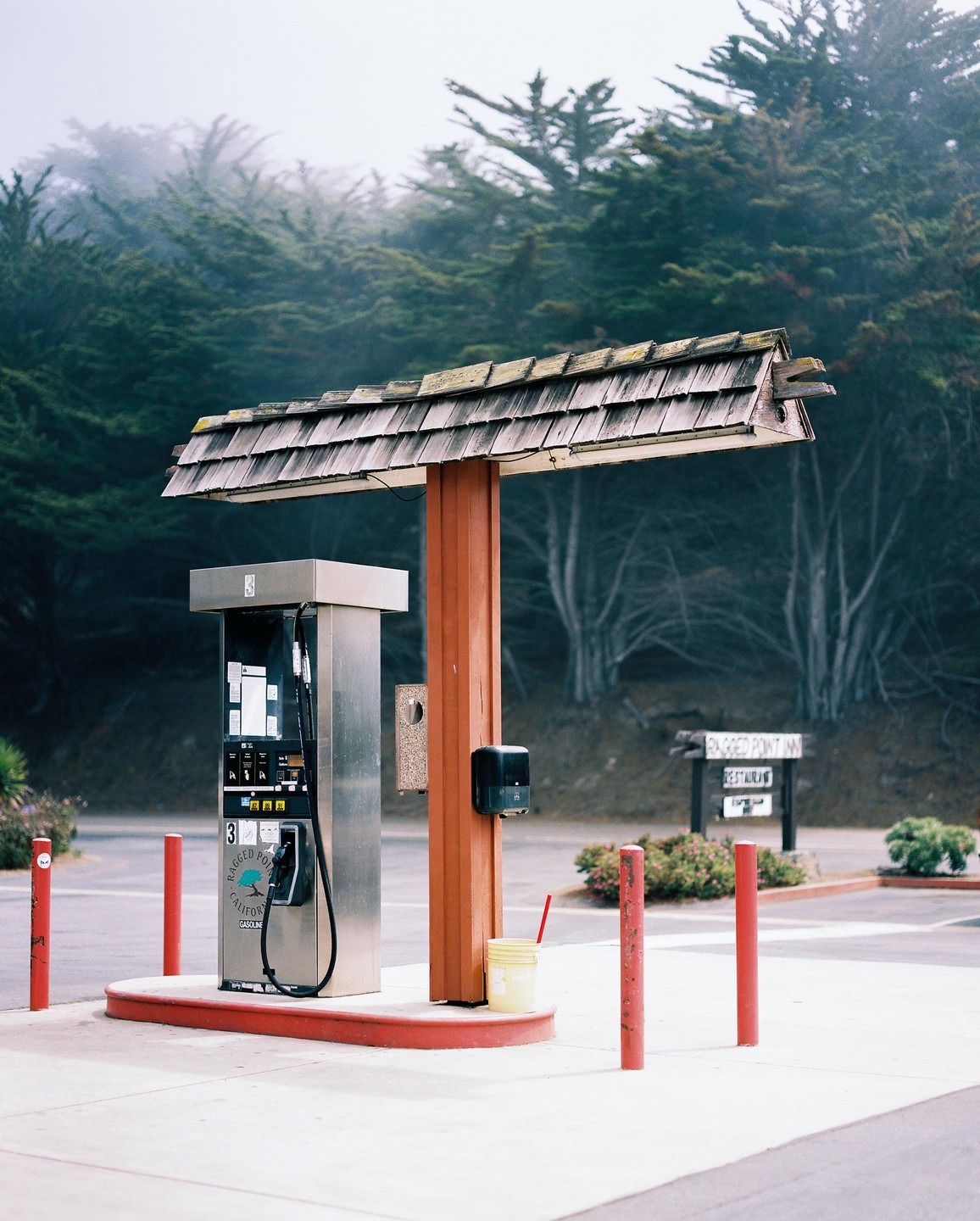 A gas pump with a wooden roof stands in a parking lot surrounded by trees and fog.