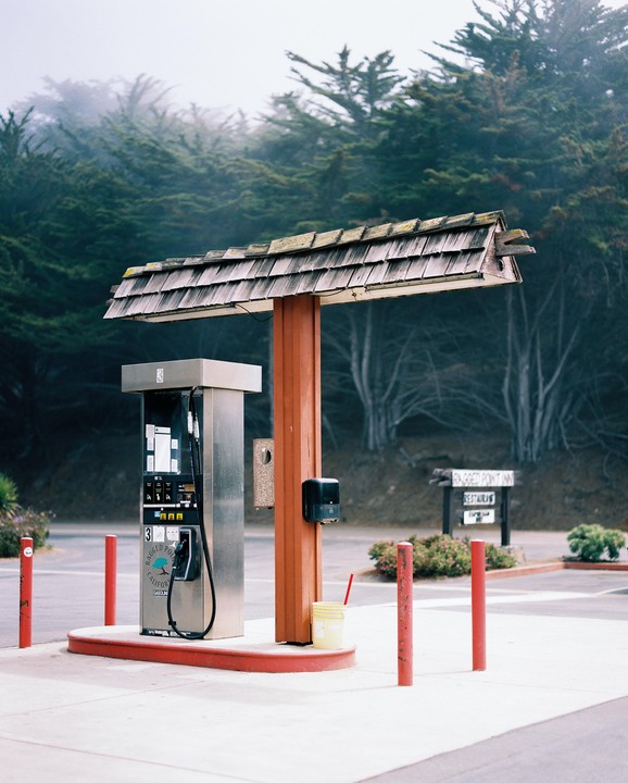 A gas pump with a wooden roof stands in a parking lot surrounded by trees and fog.