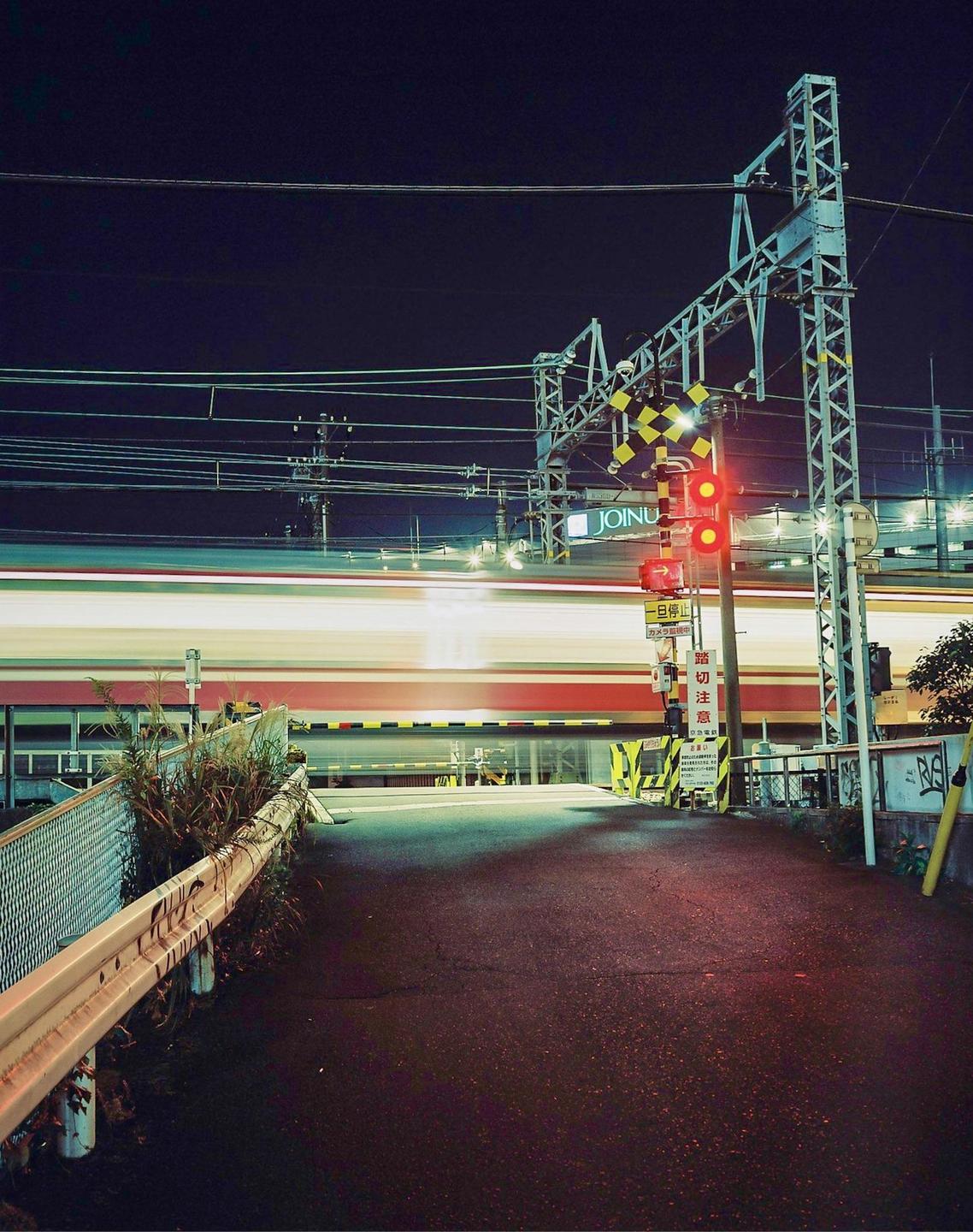 A long exposure captures the light trails of a train passing a railway crossing at night.