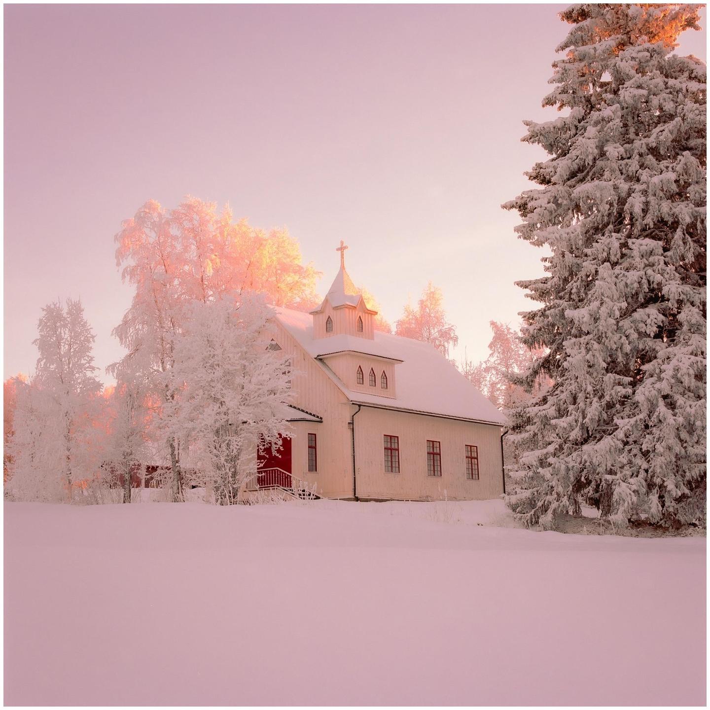 A white church with a red door and red window frames stands in a snow-covered landscape with trees.