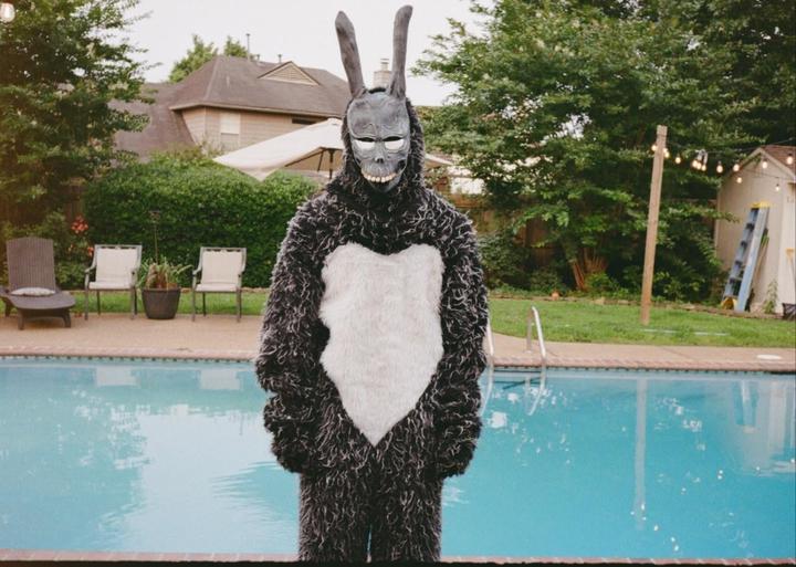 A person in a rabbit costume with a skull-like mask stands by a swimming pool in a backyard.