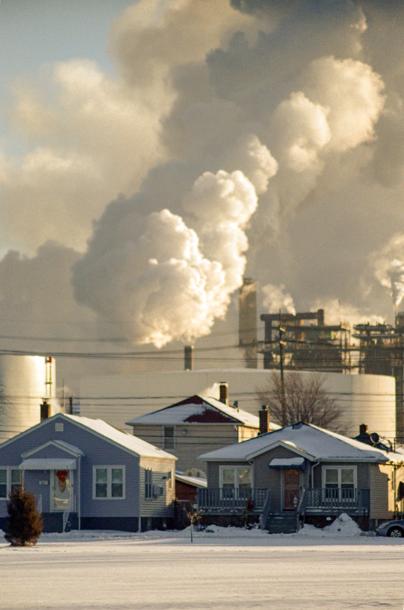 Smoke billows from an industrial plant behind snow-covered houses.