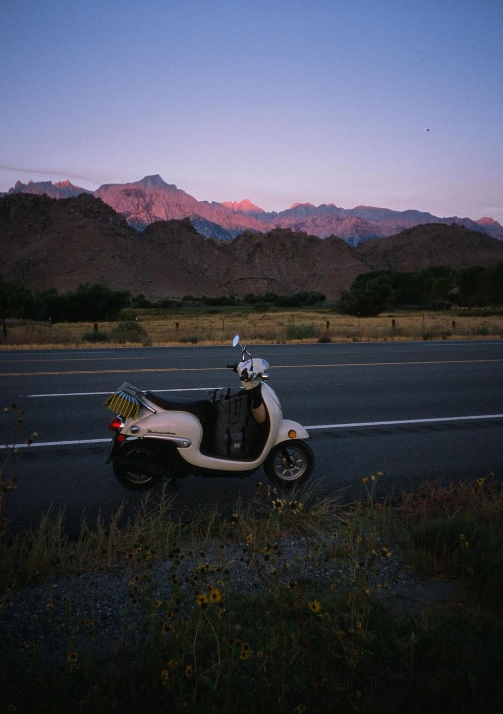 A scooter is parked on the side of a road with mountains in the background illuminated by the setting sun.