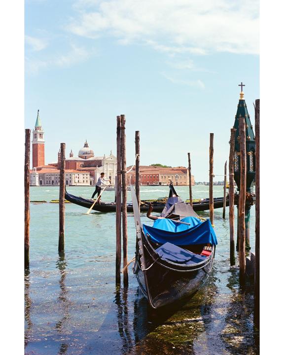 A gondola is moored to wooden posts in the foreground, with a gondolier standing in another gondola in the background, and a church and other buildings across the water.