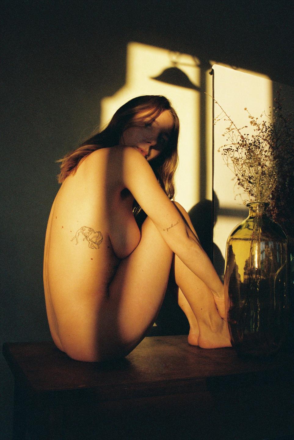 A nude woman with long hair sits on a wooden surface next to a glass vase with dried flowers, illuminated by warm light and shadow.
