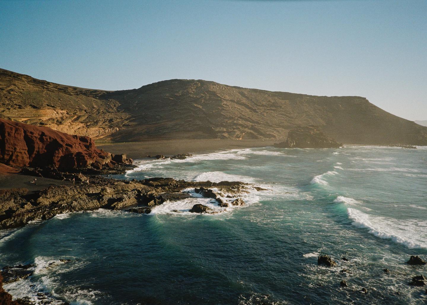 Waves crash against a rocky coastline with a dark sand beach and mountains in the background.