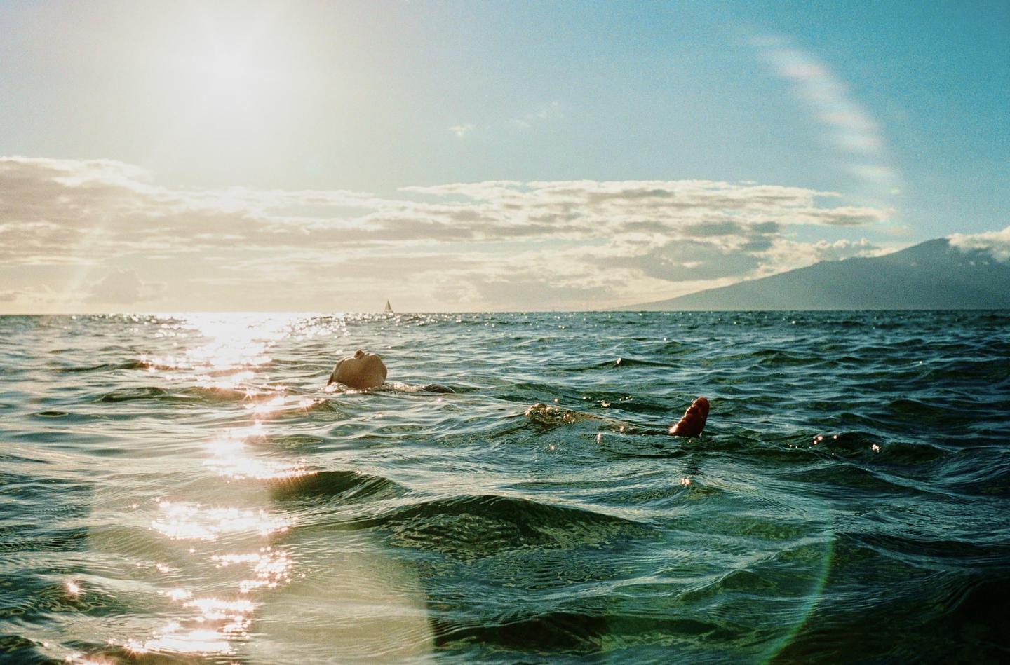 A person floats on their back in the ocean with a mountain in the distance under a bright sky.
