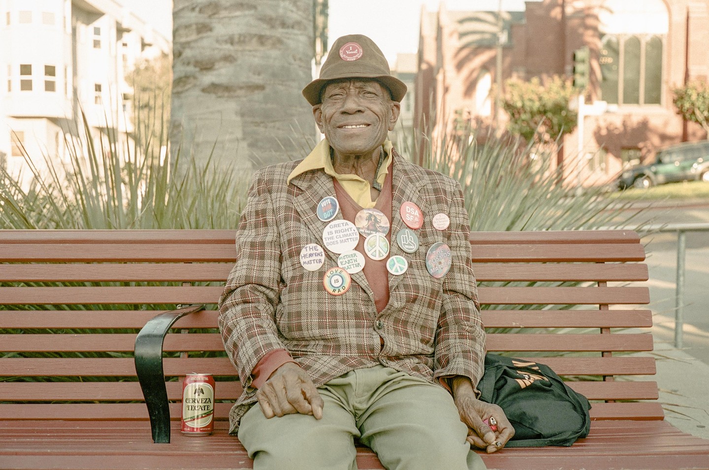 A smiling man wearing a plaid jacket covered in buttons sits on a park bench next to a can of beer.