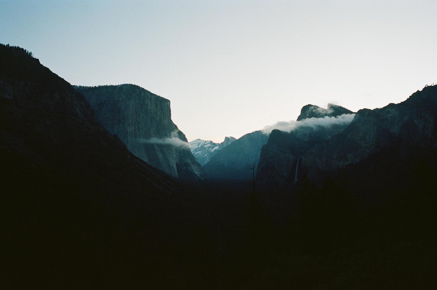 A wide valley is surrounded by tall, dark mountains with clouds clinging to their peaks and a waterfall visible on the right.