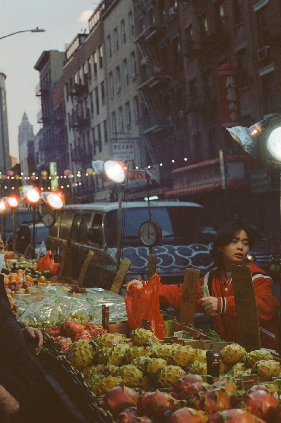 A woman in a red jacket stands behind a fruit stall on a street in new york.