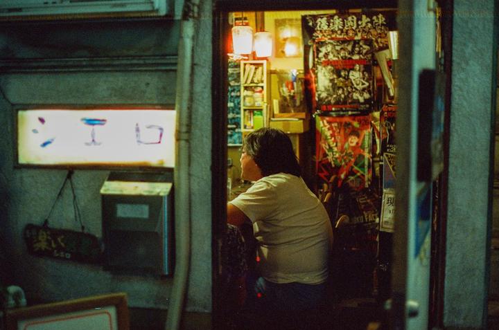 A person sits inside a brightly lit establishment with posters on the wall, viewed from a dark street at night.