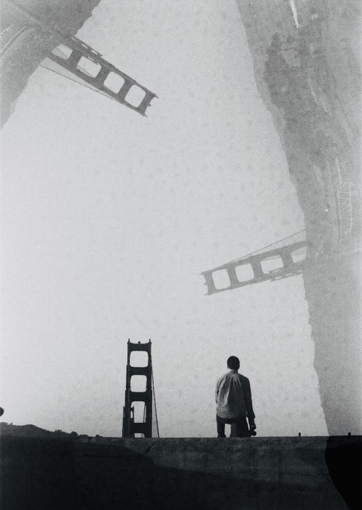 A man stands on a ledge looking towards a bridge tower, with multiple exposures of bridge structures in the sky.