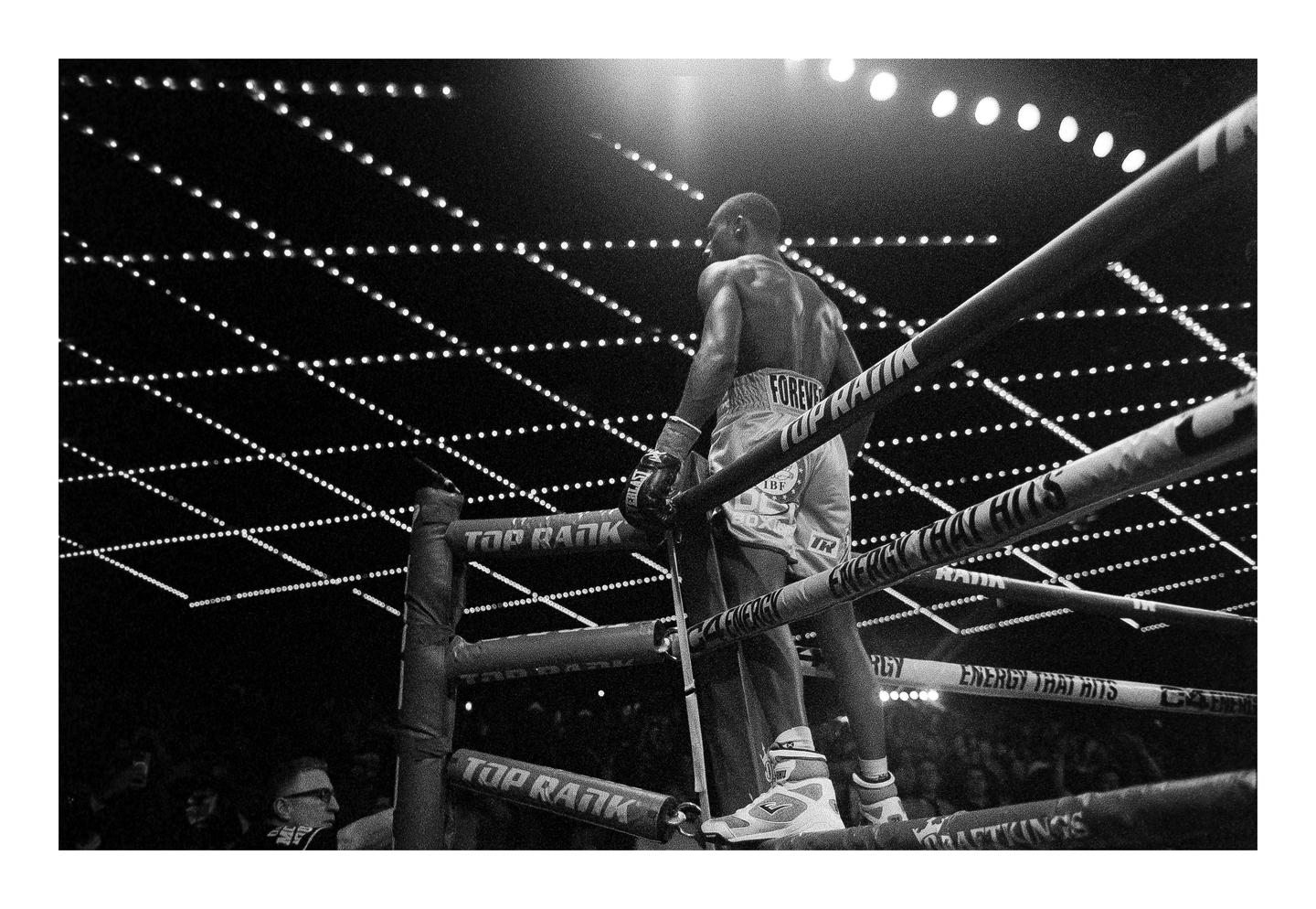 A boxer stands in a boxing ring, looking out at the crowd and the illuminated ceiling.
