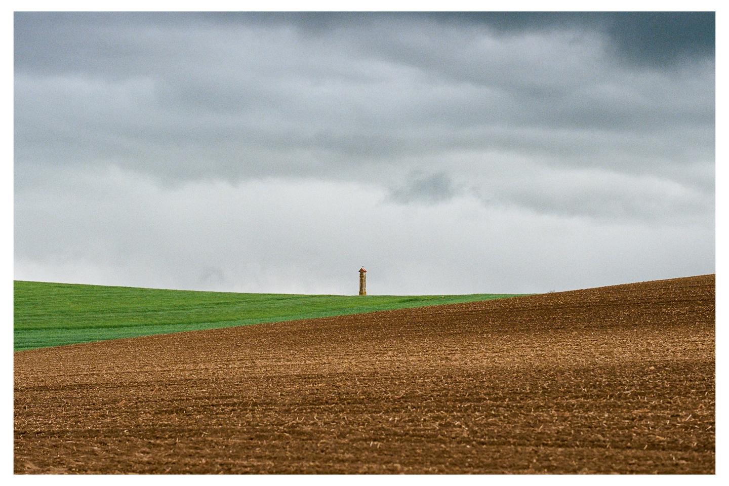 A small tower stands on a green hill under a cloudy sky, with a brown field in the foreground.