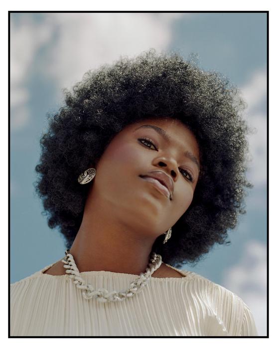 A woman with an afro and a white necklace looks up at the camera against a blue sky with clouds.