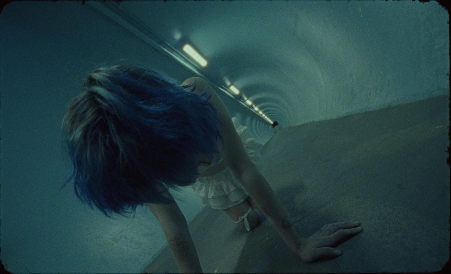 A woman with blue hair crawls on a concrete surface in a tunnel.