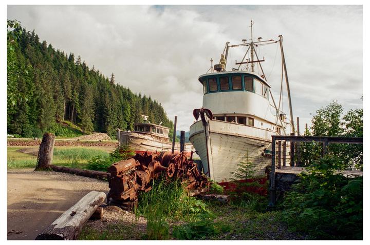 Two old fishing boats are beached on a grassy shore next to a forest under a cloudy sky.