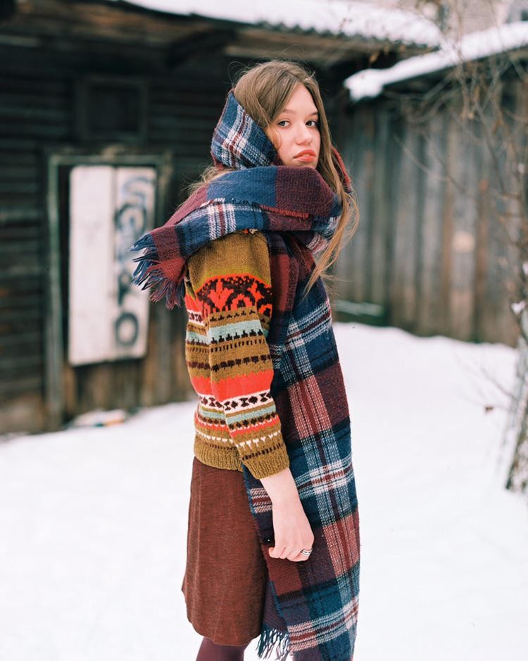 A woman wearing a colorful sweater and a plaid scarf stands in the snow in front of a wooden building.