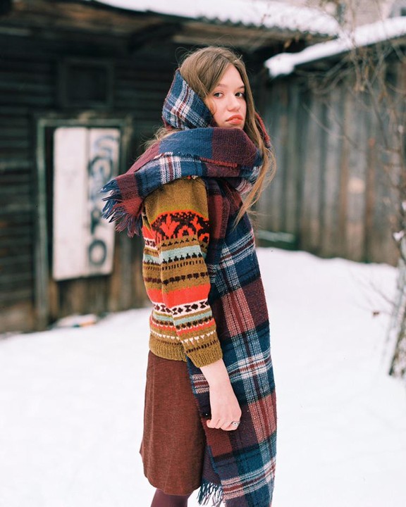 A woman wearing a colorful sweater and a plaid scarf stands in the snow in front of a wooden building.