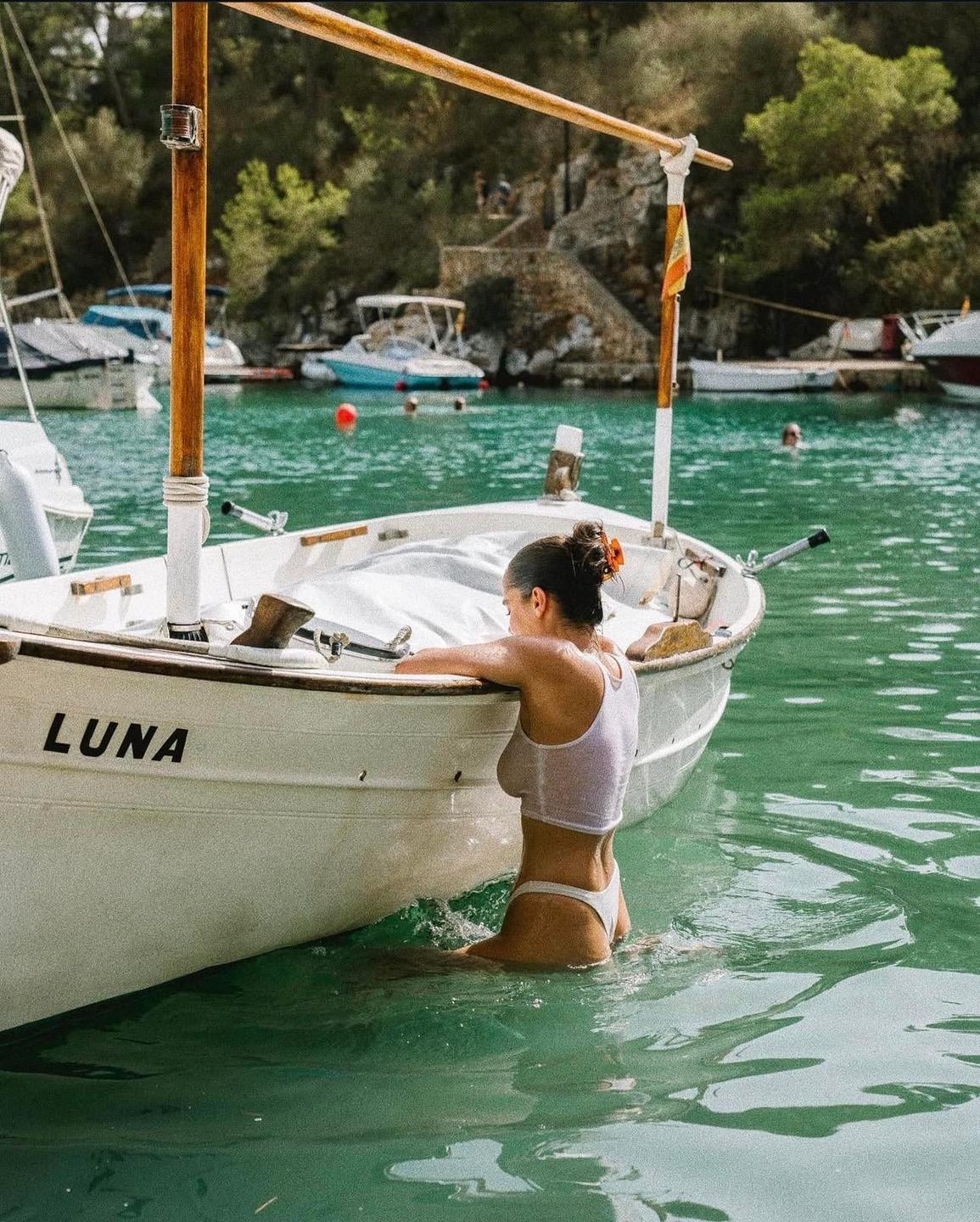 A woman in a white top and bikini bottom is in the water next to a small boat named Luna.