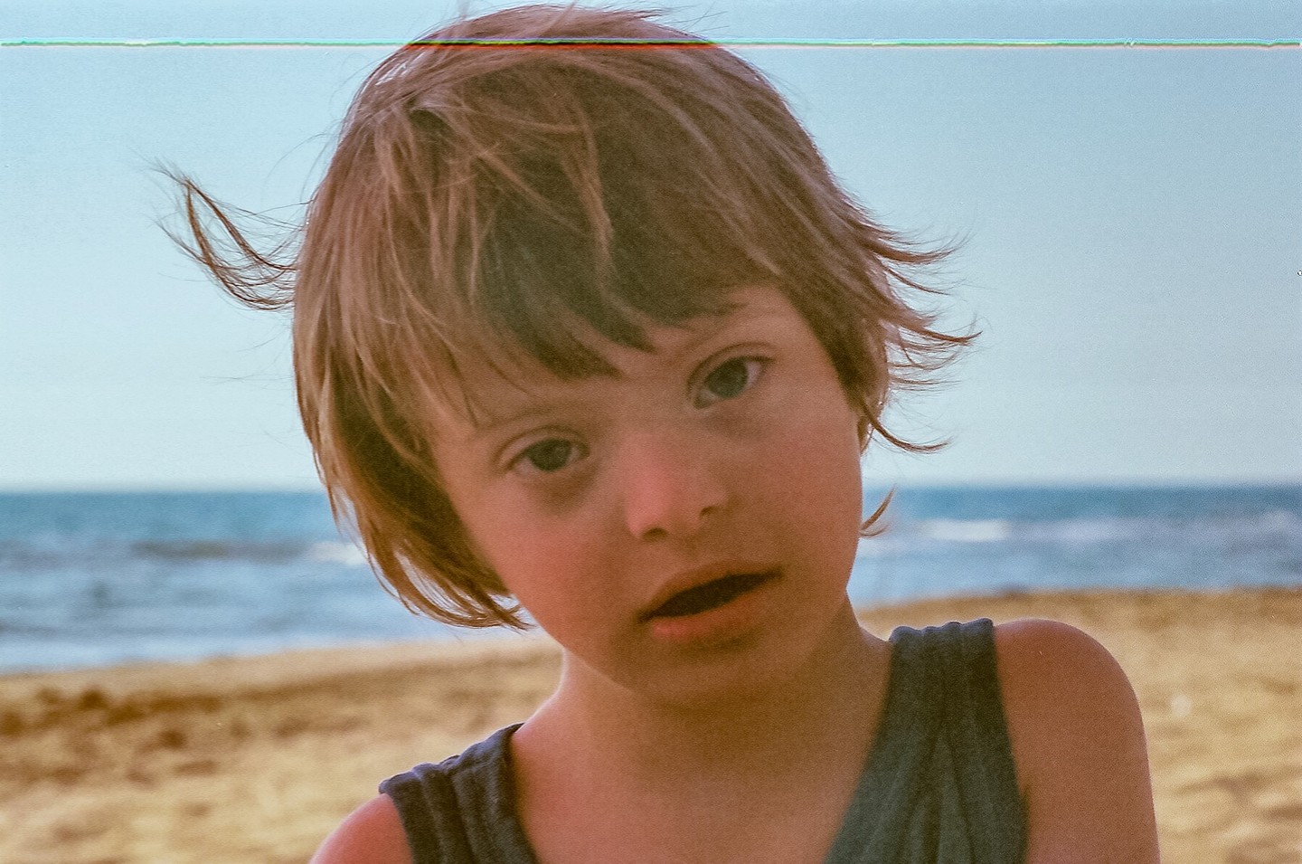 A boy with short hair looks at the viewer from a beach with the ocean in the background.