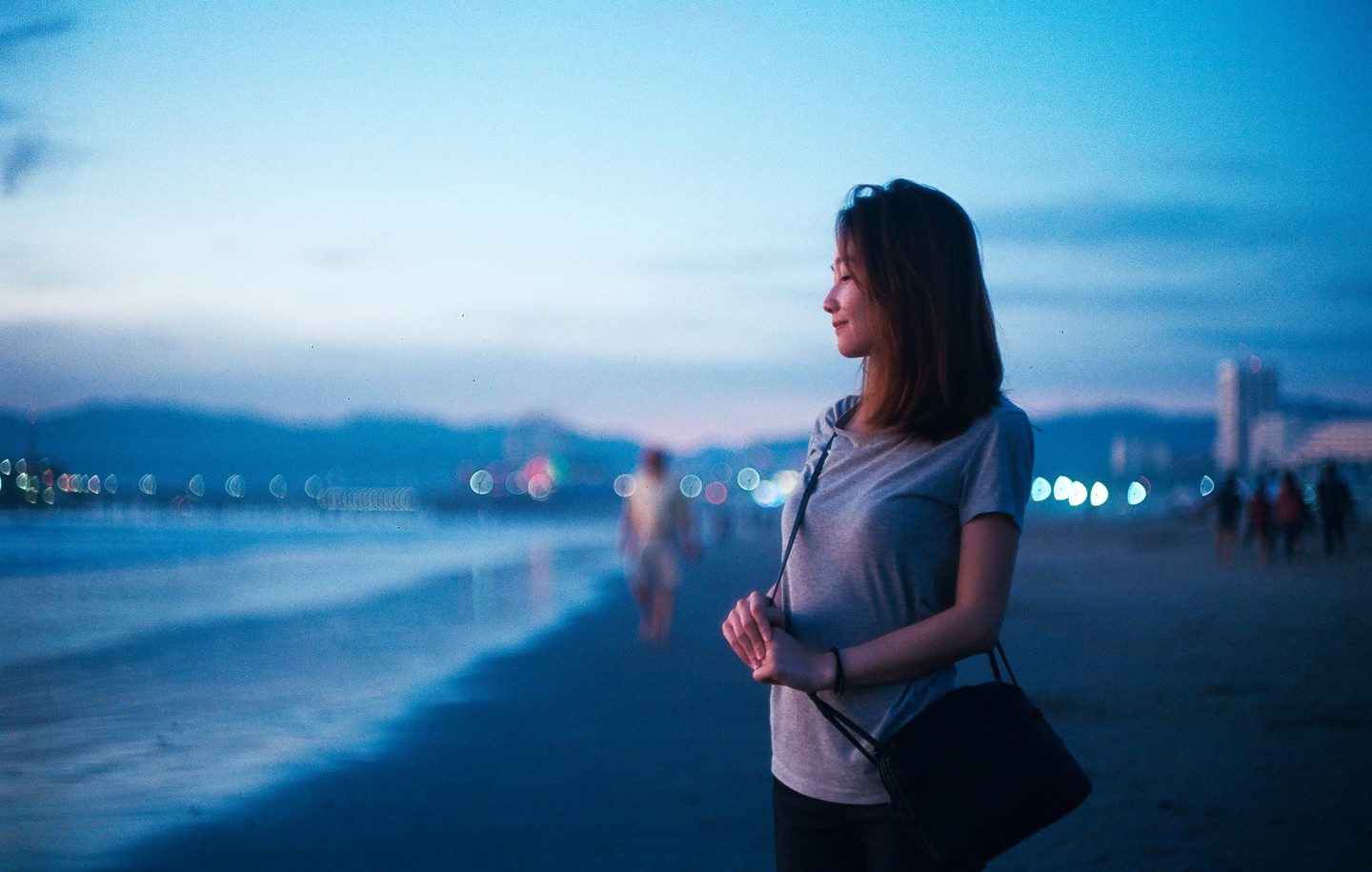 A woman stands on a beach at dusk, looking towards the water with city lights in the background.