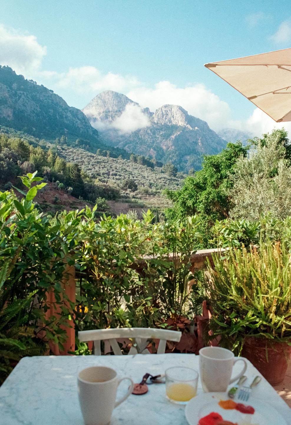A breakfast table with cups, juice, and fruit is set on a balcony overlooking a mountainous landscape with clouds.