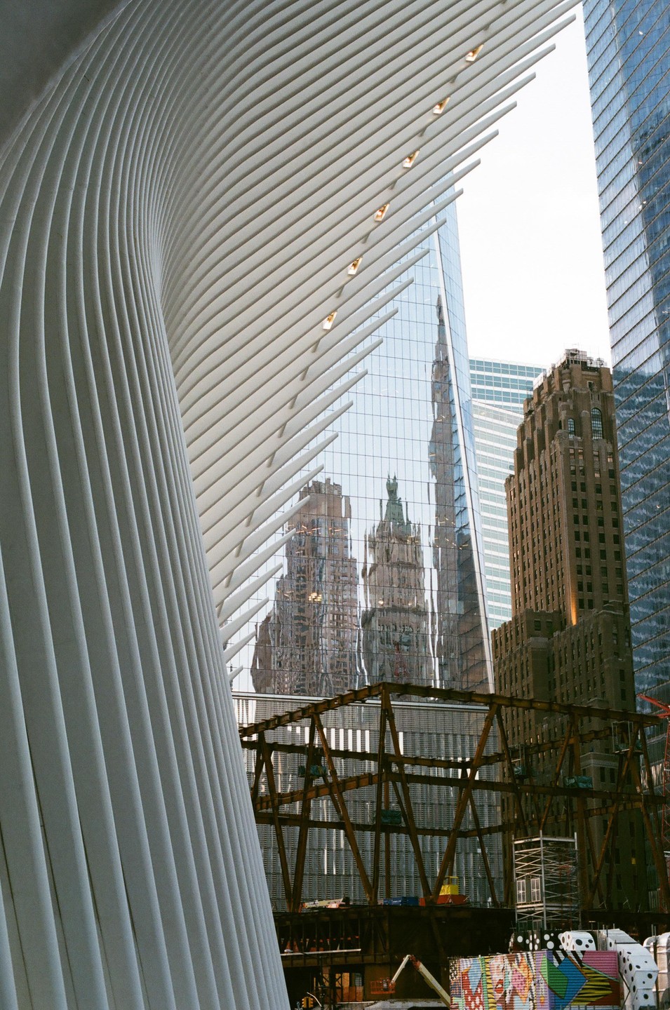 A modern white building with a ribbed facade curves around a glass skyscraper reflecting older buildings, with a brown building and construction visible in the foreground.