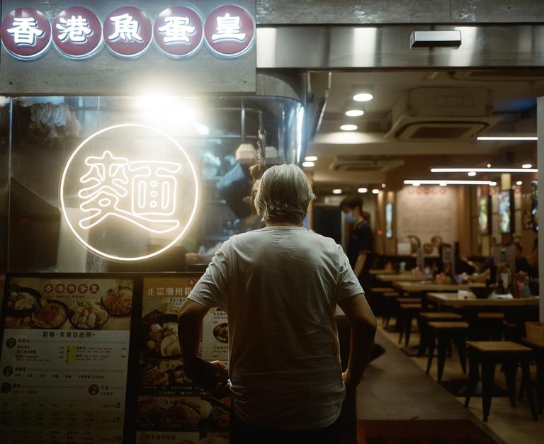 A man stands with his back to the camera, looking at a neon sign and menu outside a restaurant.