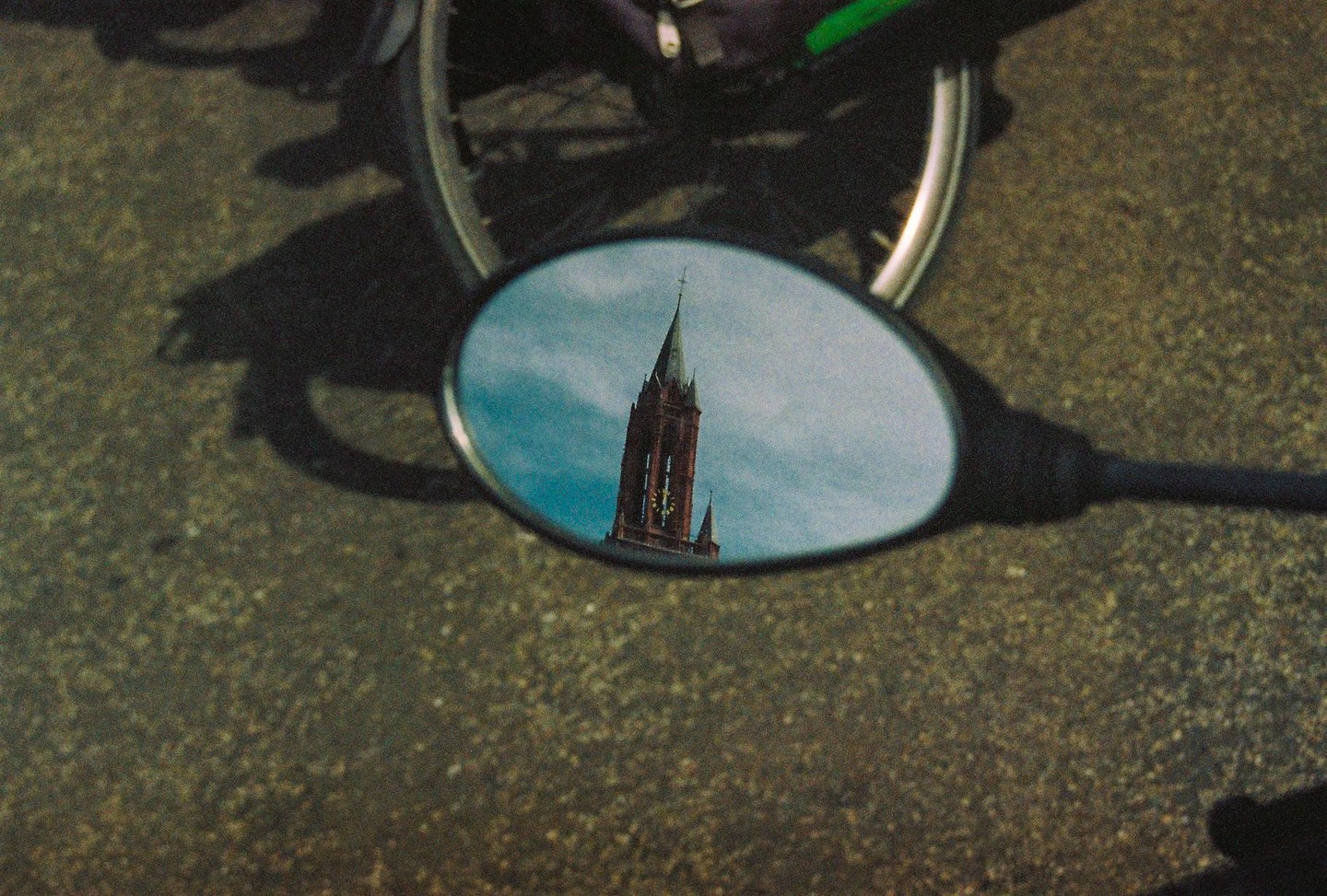 A church tower is reflected in a motorcycle mirror on an asphalt road.