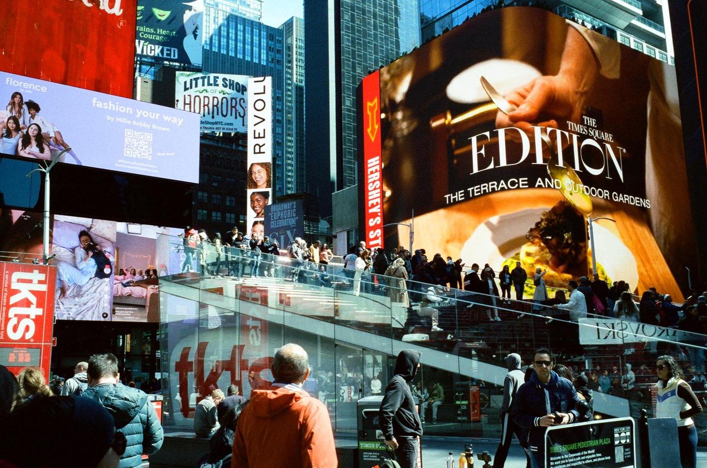A crowd of people stands on a pedestrian plaza surrounded by large illuminated billboards in Times Square.