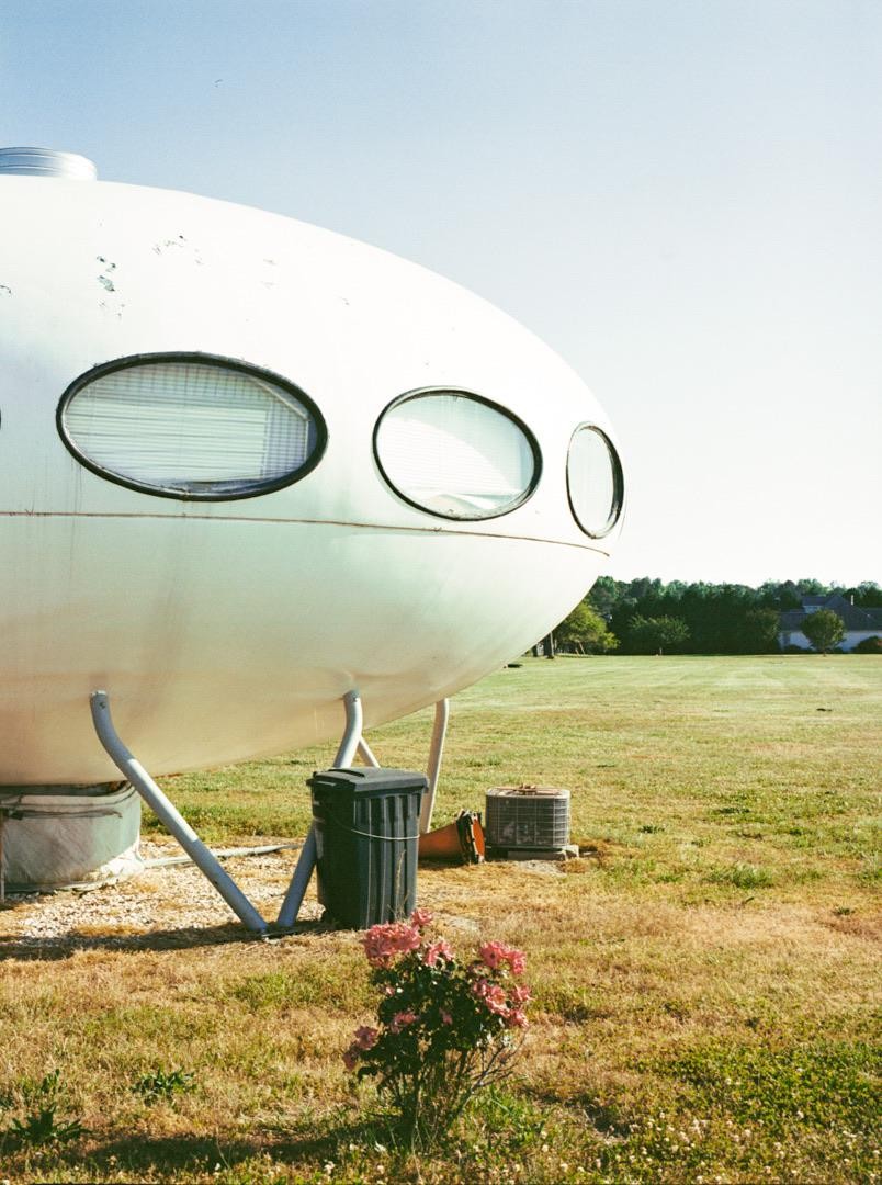 A white Futuro House with oval windows stands in a grassy field under a clear sky.