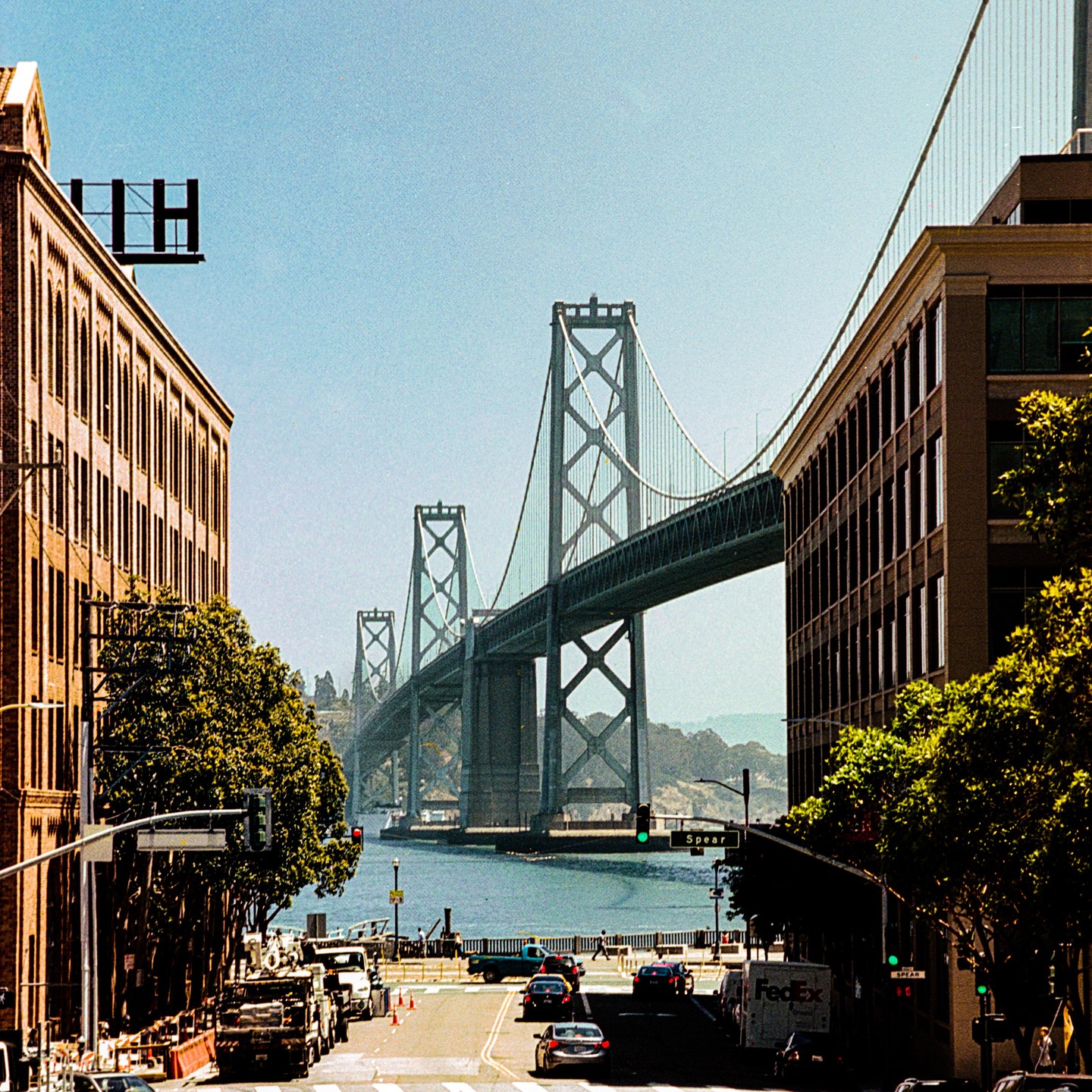 A street with cars and buildings leads to a large suspension bridge over water.
