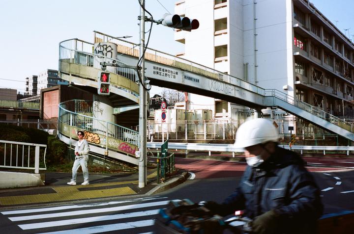 A man on a bicycle rides past a woman standing on a crosswalk next to a spiral pedestrian bridge in a city.