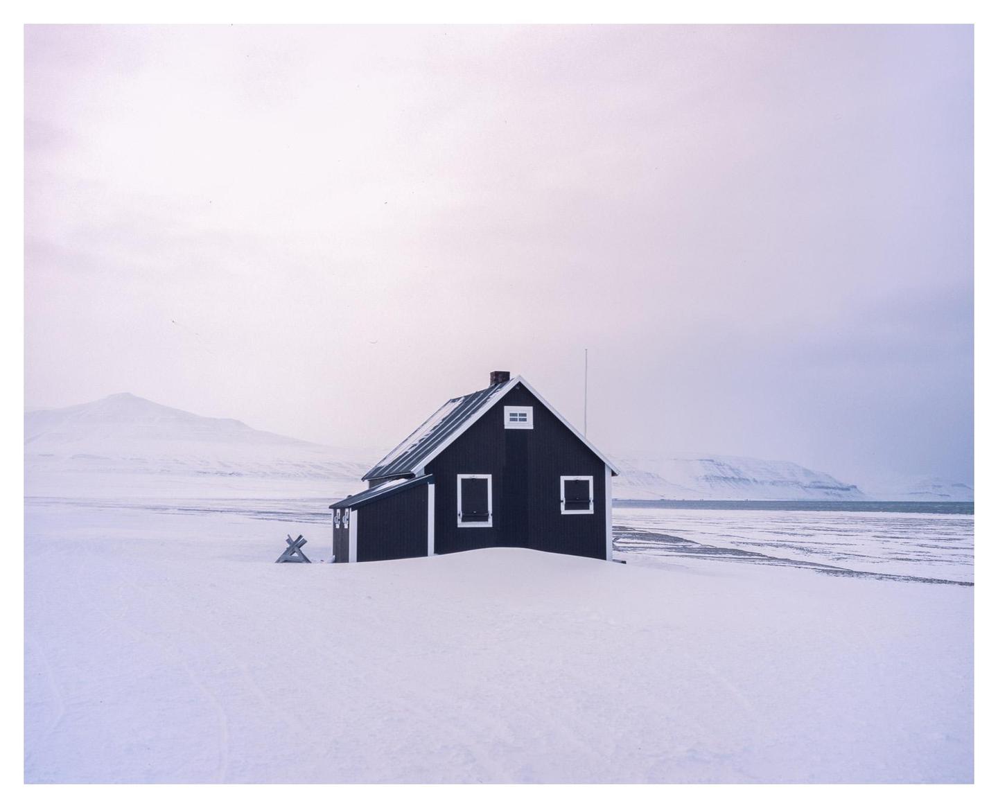 A dark house with white window frames stands in a snowy landscape next to a body of water with mountains in the background.