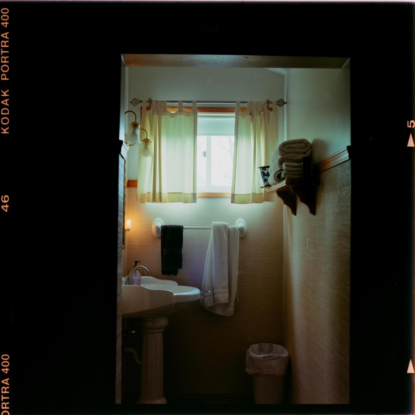 A bathroom with a pedestal sink, a window with curtains, and towels on a rack and shelf is illuminated by natural light and sconces.