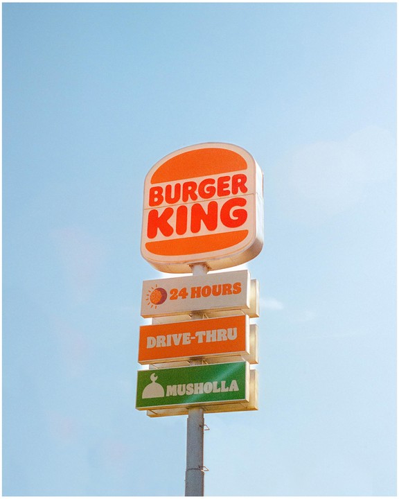 A Burger King sign with additional signs for 24 hours, drive-thru, and musholla stands against a clear blue sky.